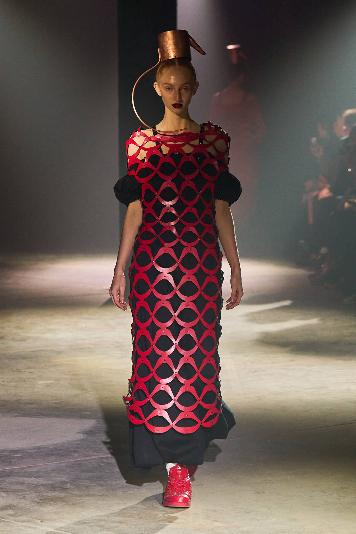 Model on a runway wearing a red circular-cutout lattice dress over black, with a copper sculptural hat and red sneakers under spotlight.