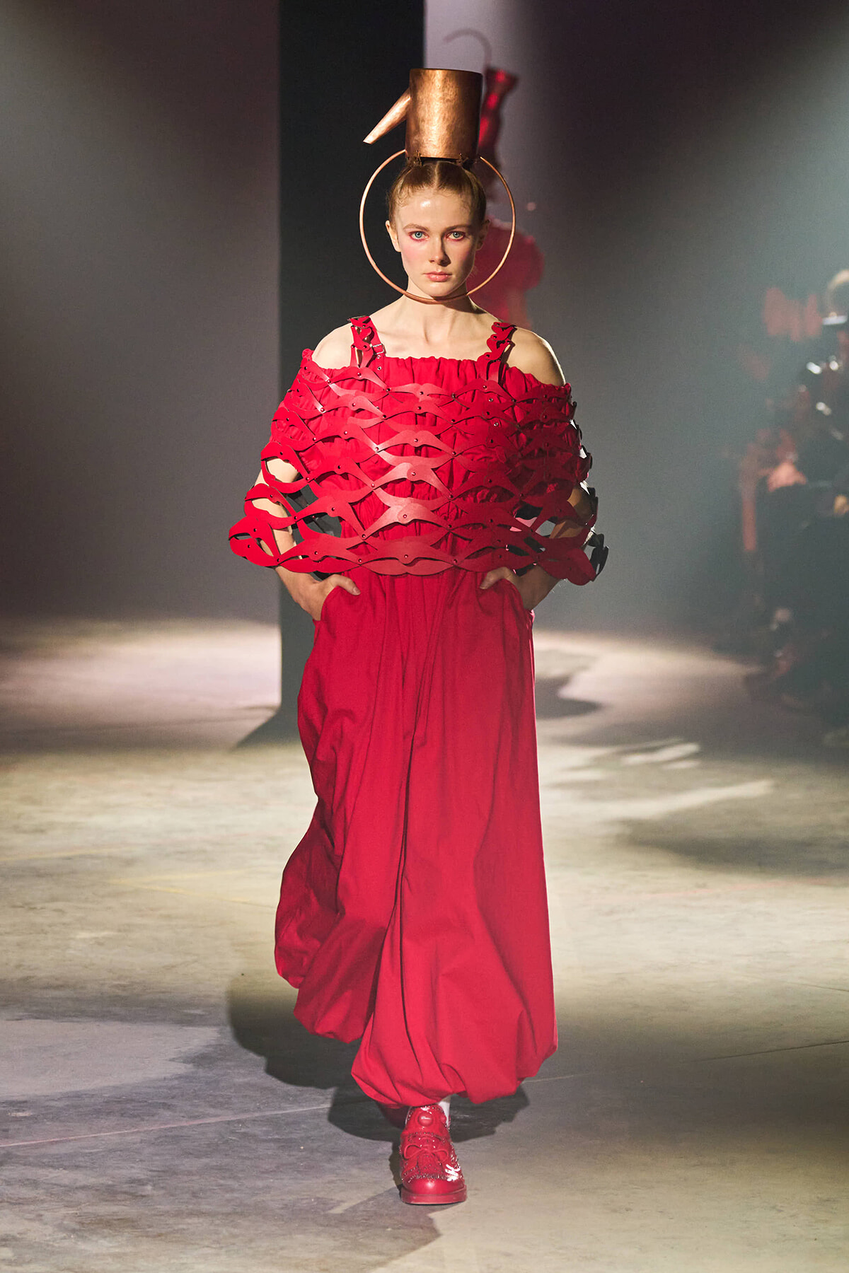Model on a runway wearing a red, voluminous gown with a lattice, cape-like upper layer and a copper top hat with large hoop earrings.