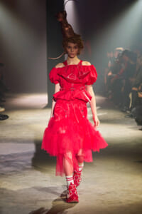 Model walking the runway in a voluminous red off-shoulder dress with a layered tulle skirt and bold red boots; copper headpiece overhead.