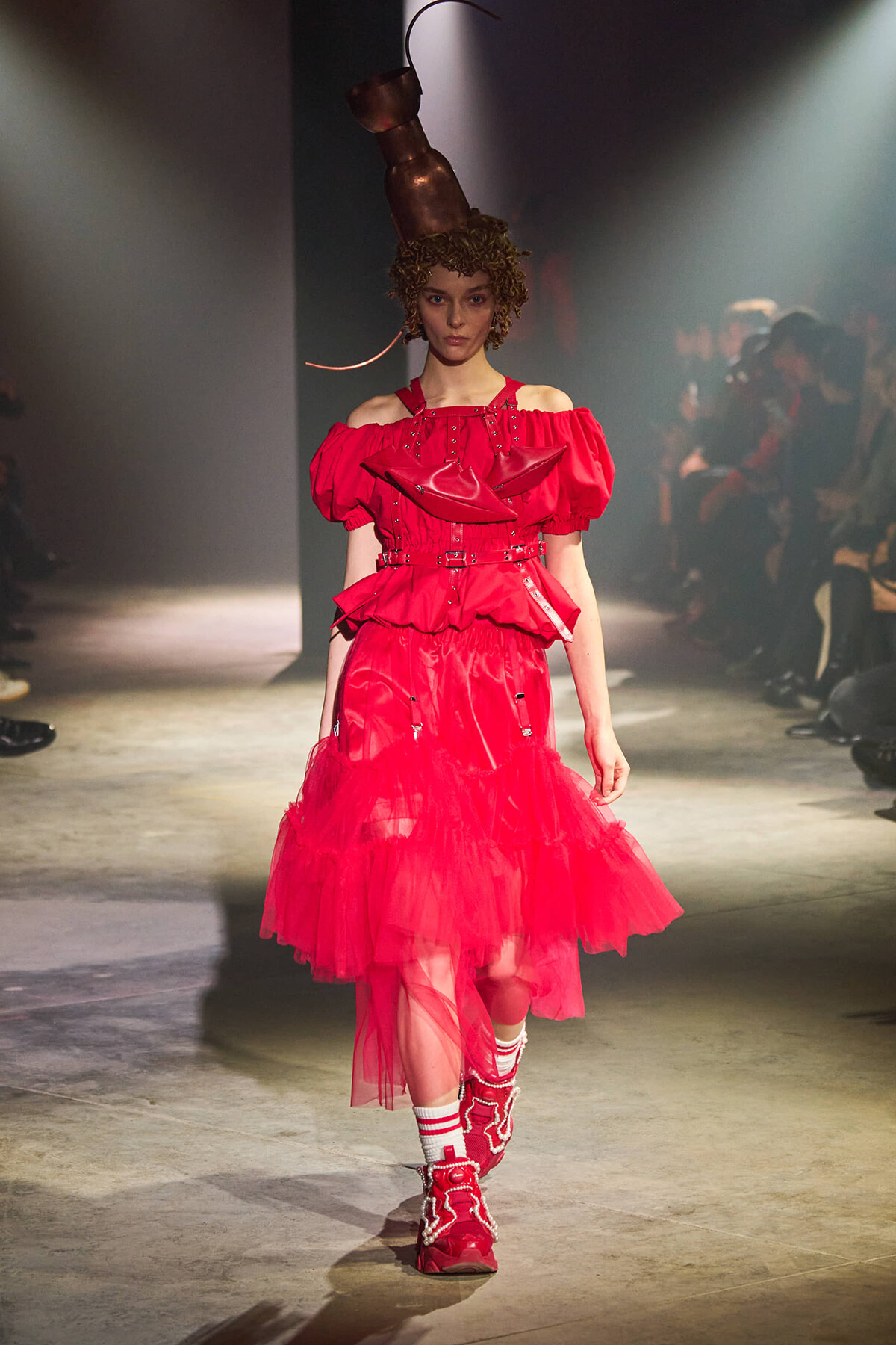 Model walking the runway in a voluminous red off-shoulder dress with a layered tulle skirt and bold red boots; copper headpiece overhead.