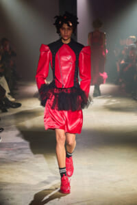 Runway model wearing a red structured jacket and matching shorts with a black corset-like harness and a black tulle waist belt.