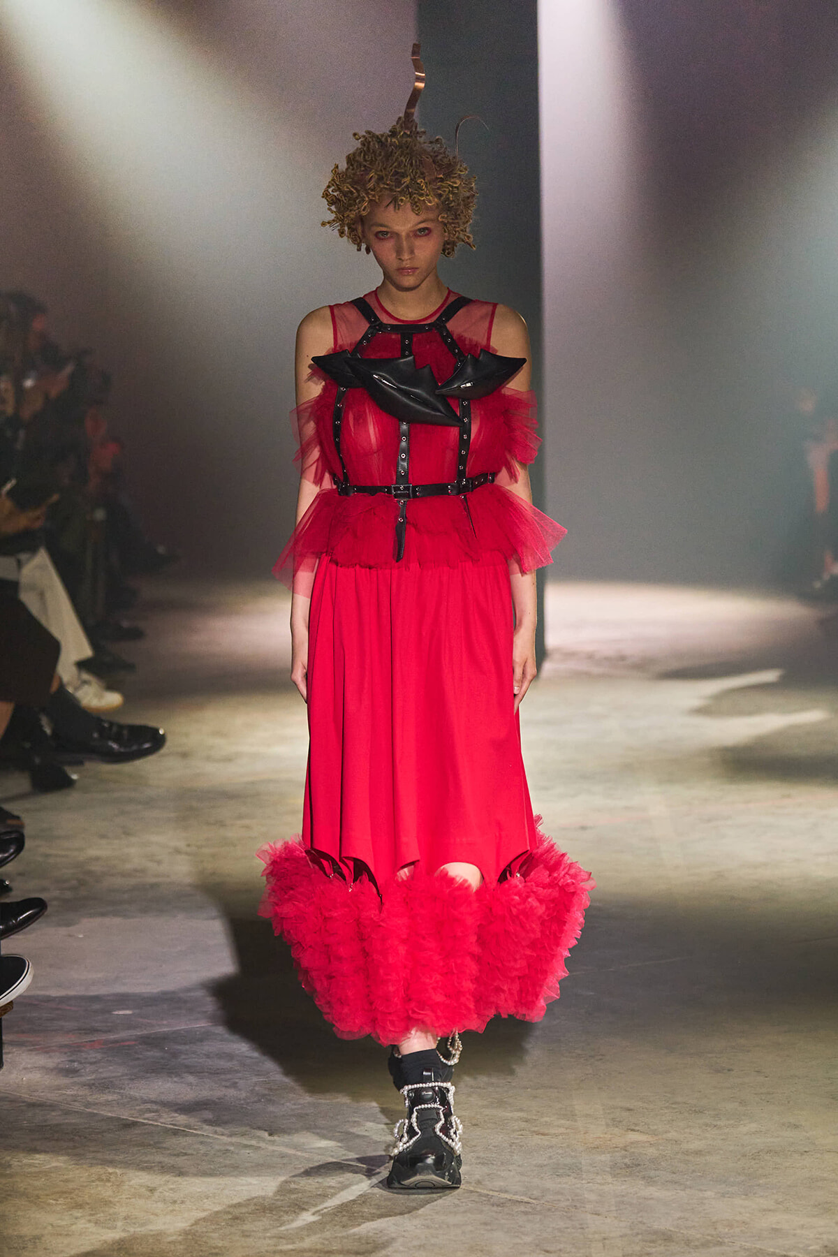 Runway model in a red haute couture dress with black harness details and a full ruffled hem, walking the catwalk.
