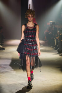 Model walks a fashion show in a black mesh dress with red lip-shaped appliqués linked by chains, layered tulle, a sculptural headpiece, pink socks, and chunky boots.