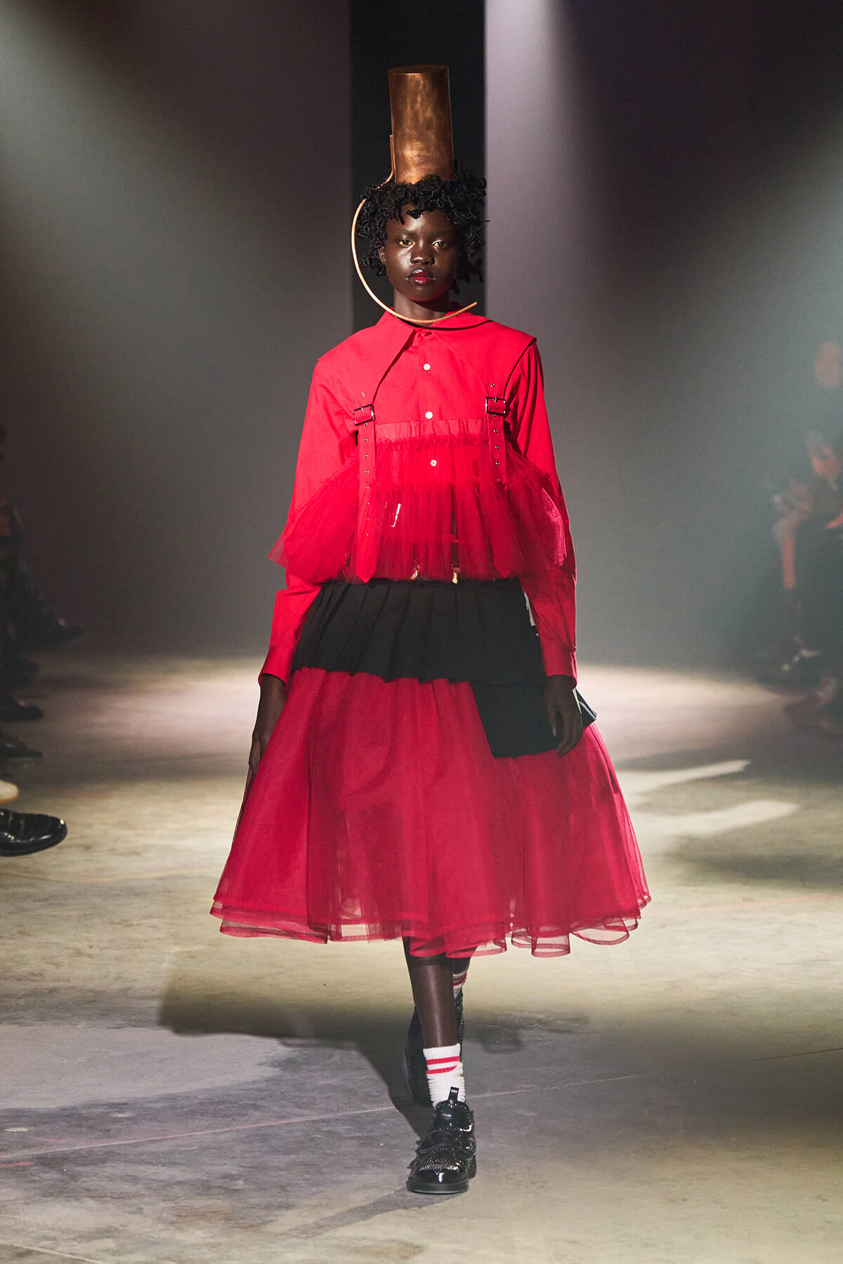 Model on a runway wearing a red sheer blouse with tulle overlay and a voluminous red skirt, black waist accents, large circular earring, and a tall cylindrical headpiece.