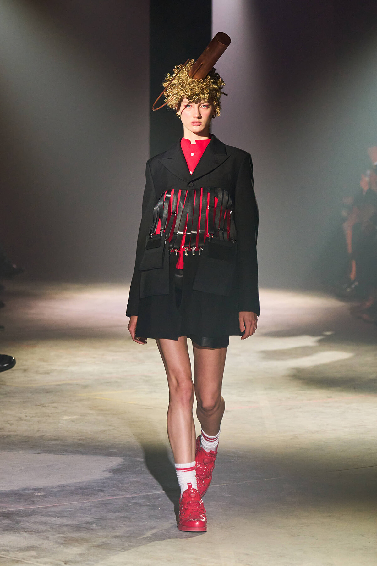 Model walks a runway in a black blazer with a red top and red strap corset, white socks, and red sneakers, topped with a gold curly wig and a large brown cylindrical headpiece.
