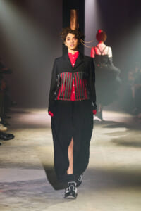 Model walks the runway in a black oversized coat with a red shirt, red gloves, and a fringe-cylinder corset detail; tall copper hat and curly hair accent the look.