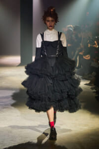 Runway model in a white button-down with a black corset-style top and a voluminous multi-layer black tulle skirt, red socks and dark boots under spotlight.