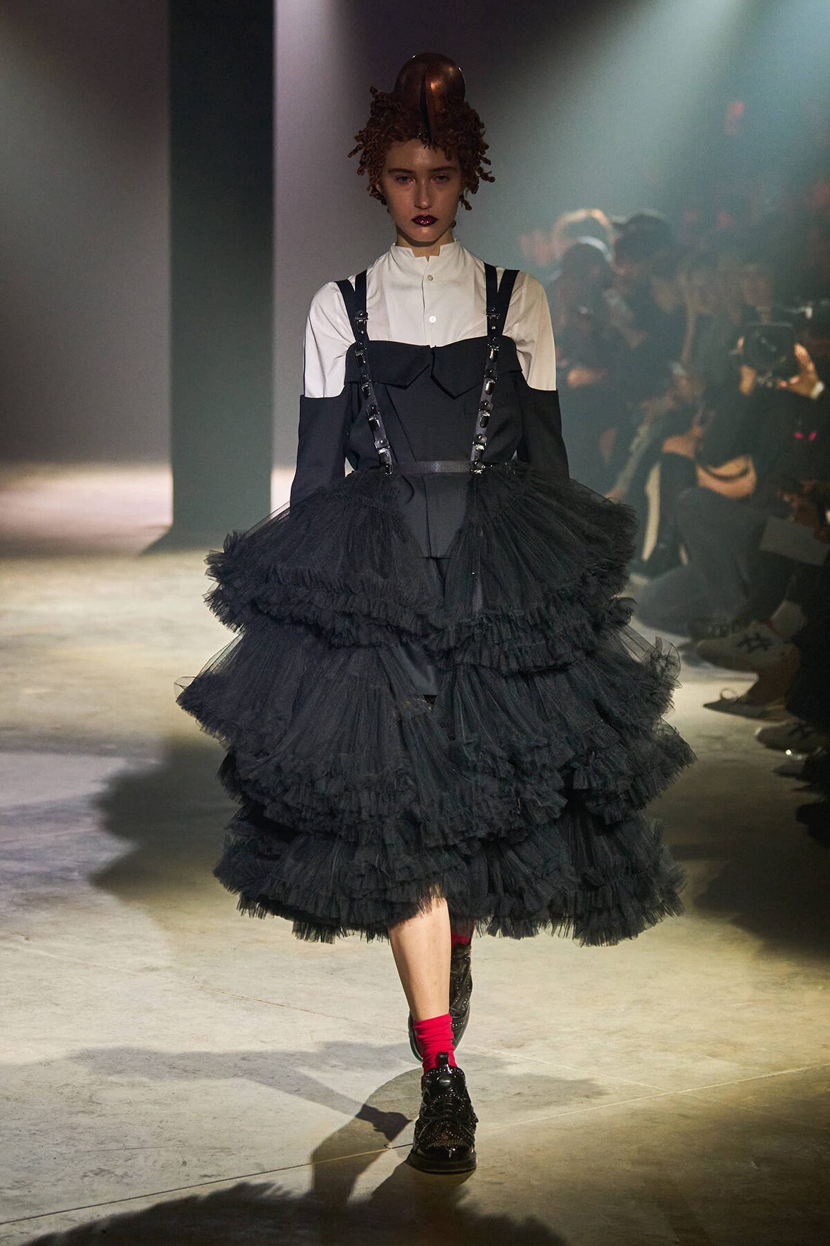 Runway model in a white button-down with a black corset-style top and a voluminous multi-layer black tulle skirt, red socks and dark boots under spotlight.