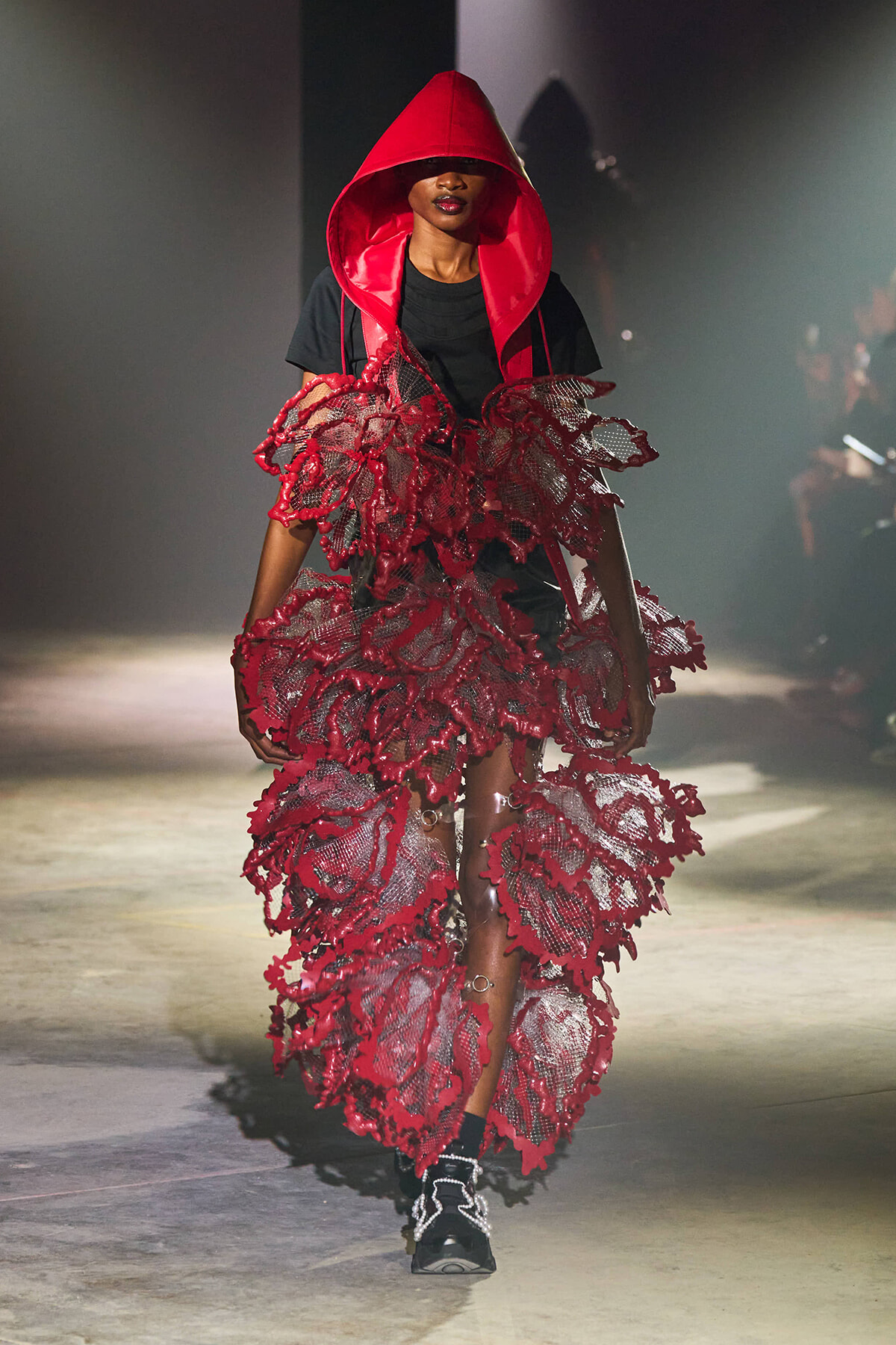 Model walks the runway in a bright red hooded cape and a dramatic red lace dress over a black outfit.