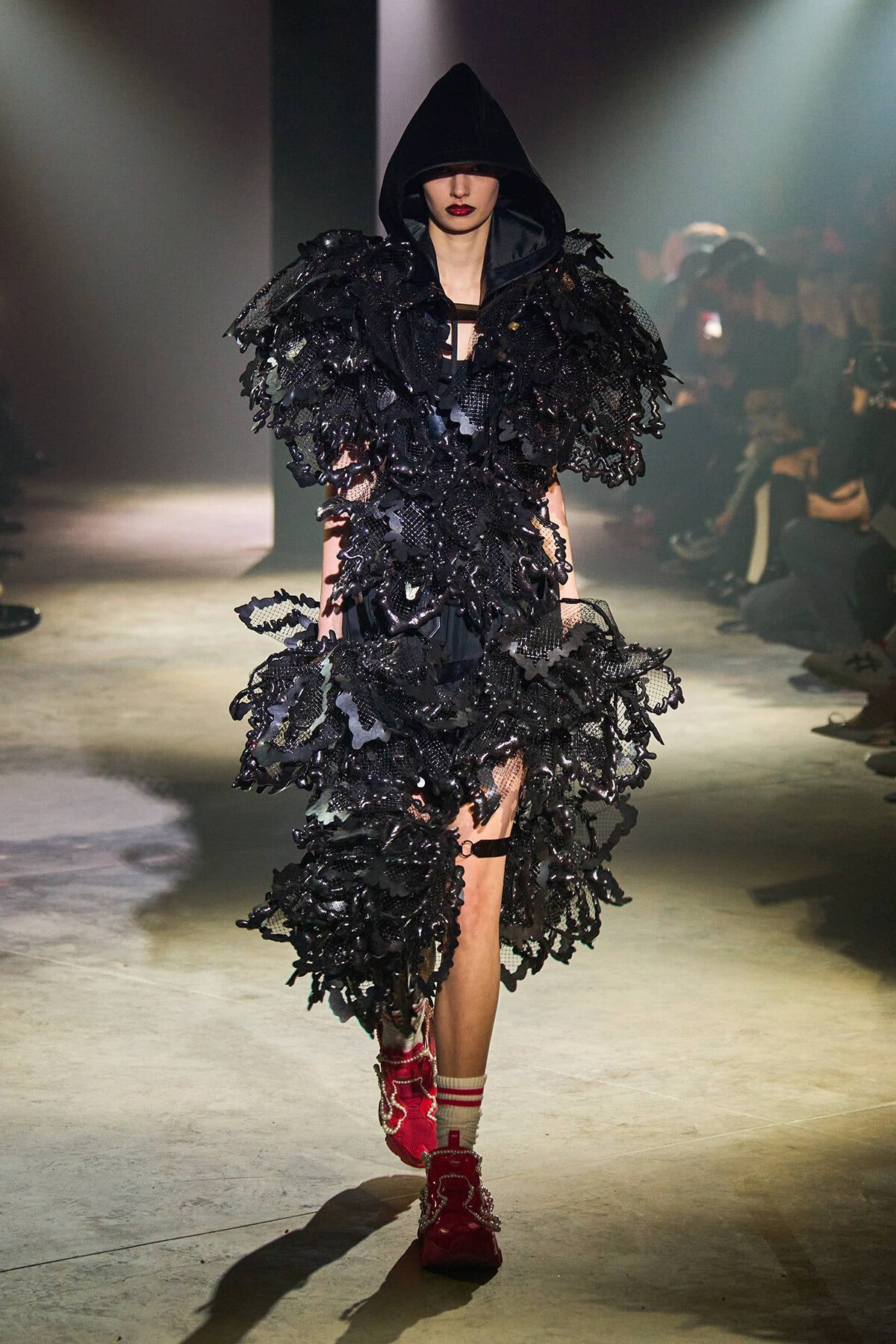 Model on a runway wearing an all-black, hooded avant-garde dress with layered leaf-like appliqués and red ankle boots with socks showing