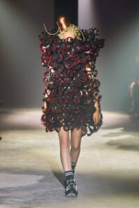 Model walking a runway in an avant‑garde dress made of dense black circular loops with red edges and a copper headpiece.