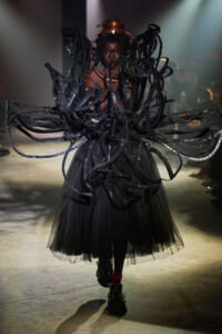 Model on a runway wearing an elaborate black couture ensemble with layered mesh wings and a tulle skirt.