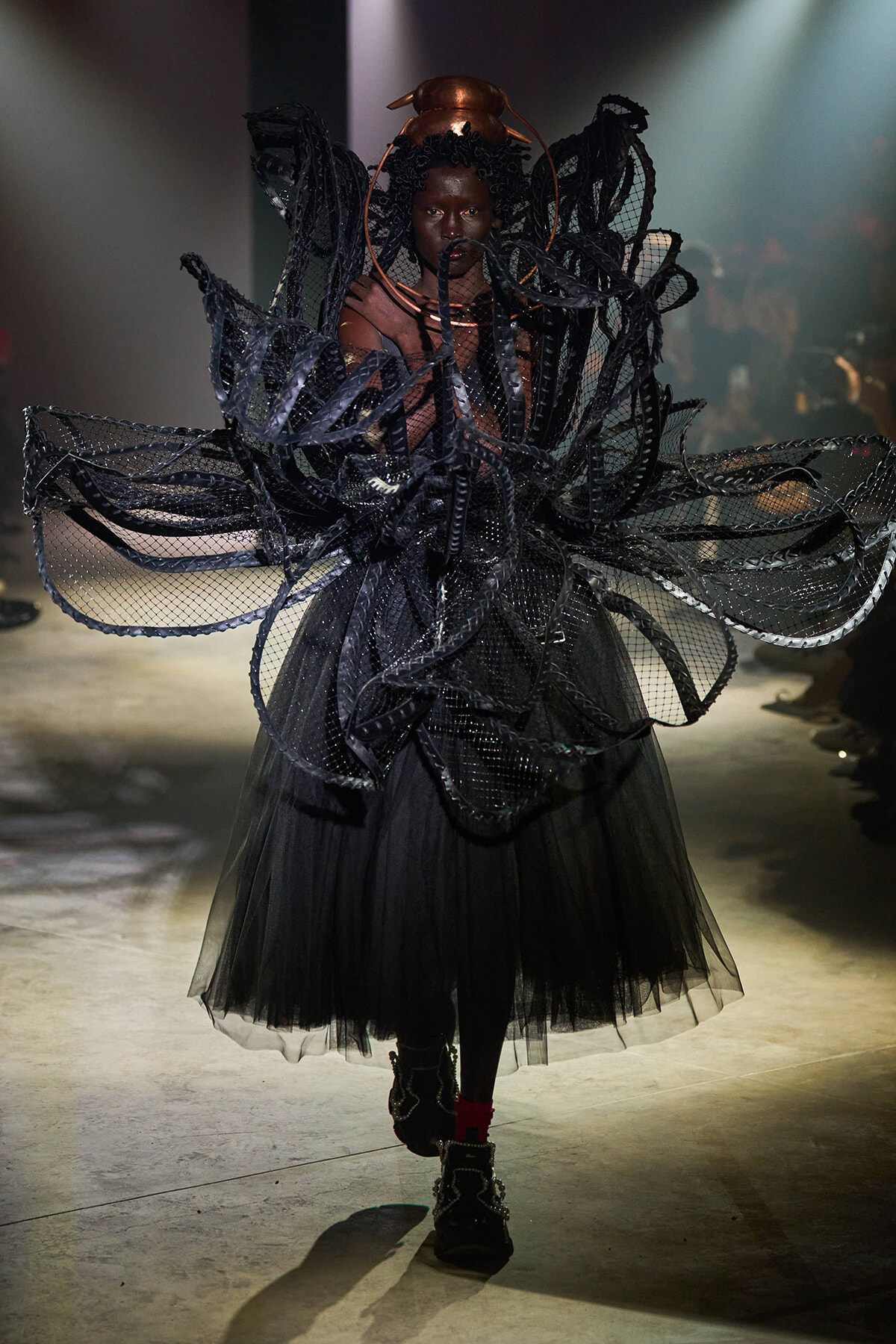 Model on a runway wearing an elaborate black couture ensemble with layered mesh wings and a tulle skirt.