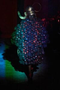 Model wearing a voluminous, bubble-like teal garment with a metallic helmet on a dark runway stage under colored lights, highlighting futuristic fashion.