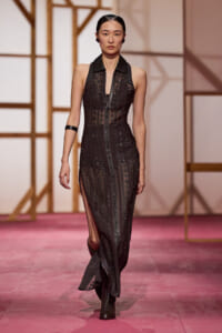 Model walks a pink runway in a sheer black geometric-pattern gown with a front zipper, sleeveless, black collar, side slits, paired with black ankle boots.