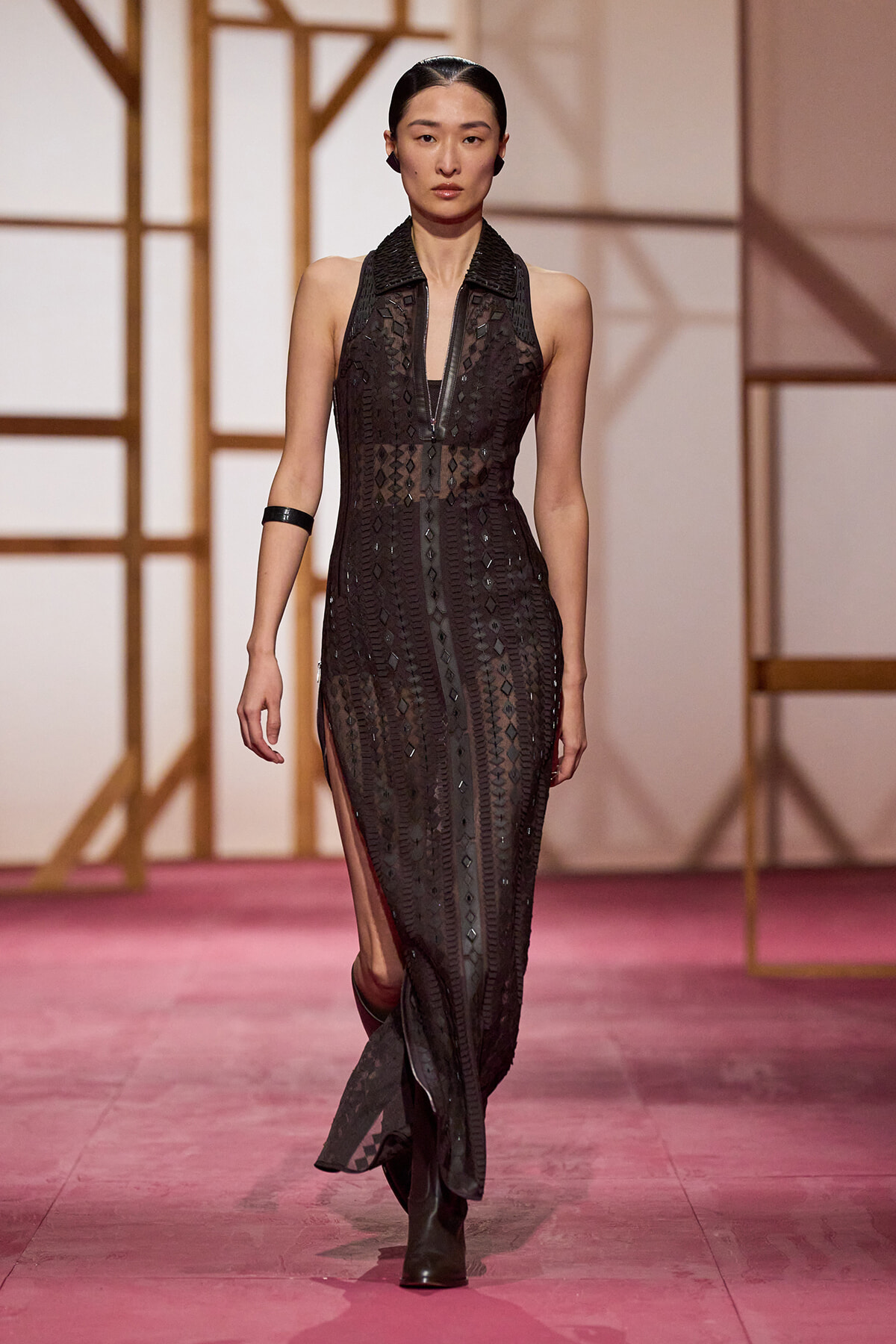 Model walks a pink runway in a sheer black geometric-pattern gown with a front zipper, sleeveless, black collar, side slits, paired with black ankle boots.