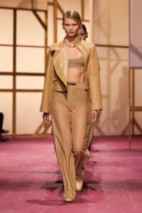 Model walking a beige monochrome outfit—crop top, wide-leg pants, and a draped jacket—on a pink runway.