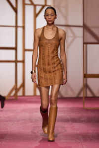 Female model walking a fashion runway in a tan geometric-patterned mini dress and knee-high tan boots, pink runway floor in view behind.