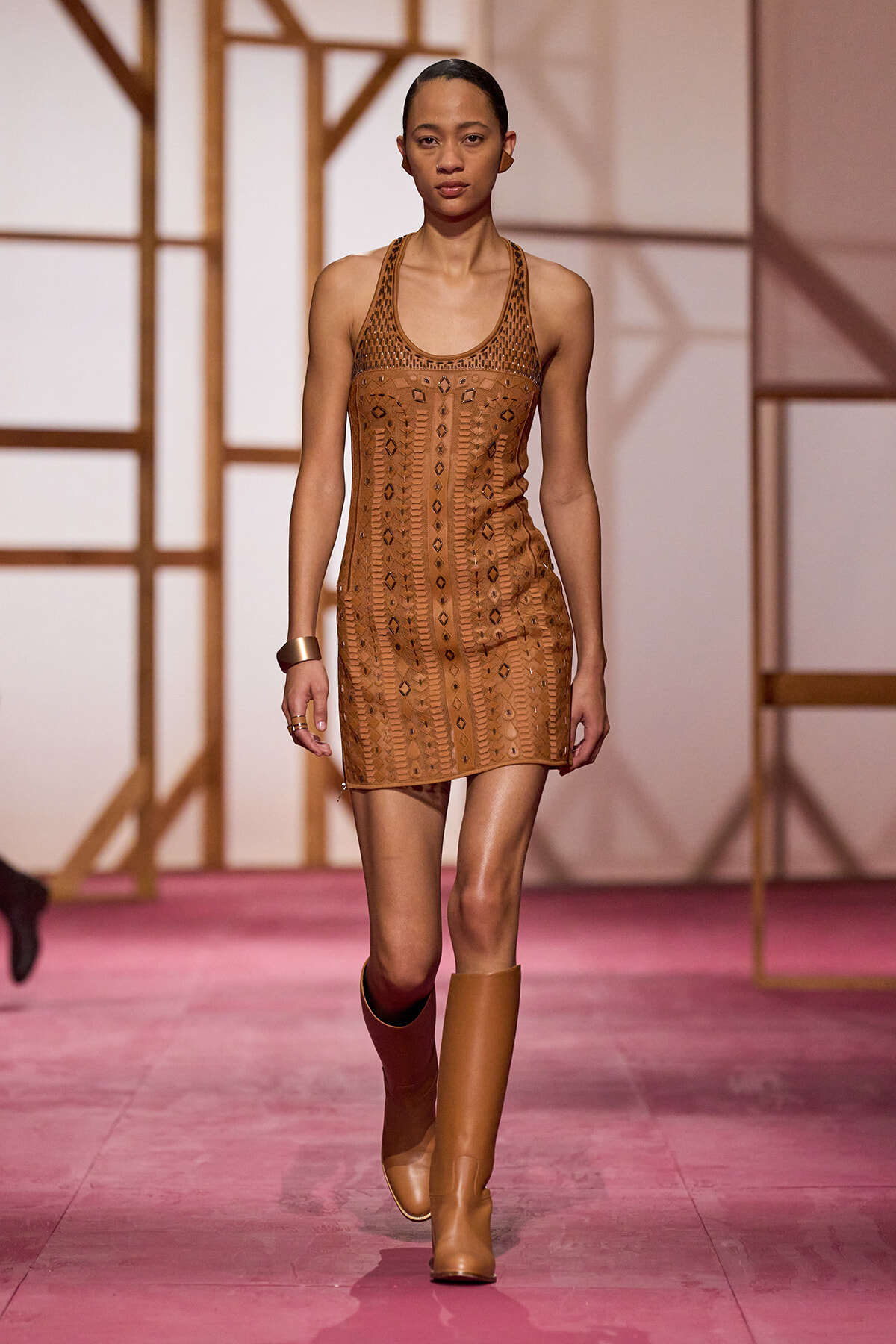 Female model walking a fashion runway in a tan geometric-patterned mini dress and knee-high tan boots, pink runway floor in view behind.