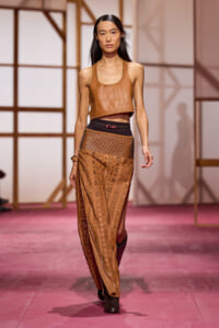 Runway model walking the catwalk in a tan cropped leather top and sheer patterned pants with a black waistband.