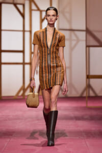 Model walking a runway in a brown mini dress with black geometric cutouts and a front zipper, paired with black knee-high boots and a woven basket handbag.
