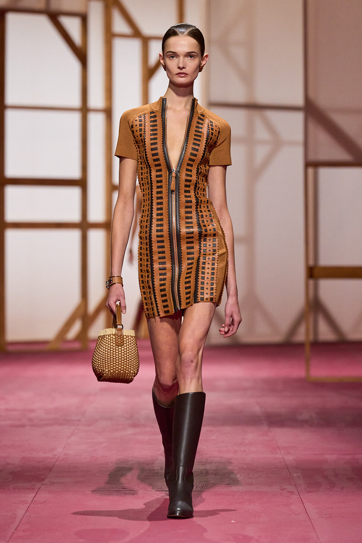 Model walking a runway in a brown mini dress with black geometric cutouts and a front zipper, paired with black knee-high boots and a woven basket handbag.