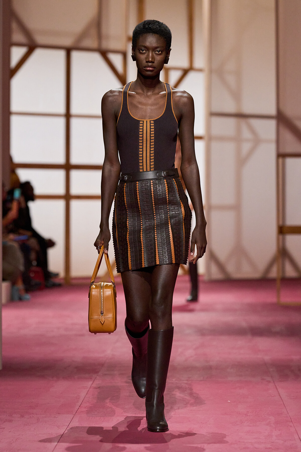Model on a pink runway wearing a brown striped knit mini dress with orange trim, black knee-high boots, and a tan handbag.