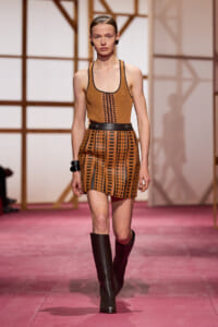 Model walking a fashion runway in a brown sleeveless top and geometric-pattern mini skirt with a black belt and tall boots.