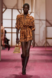 Model walks a fashion runway in a tan-brown patterned romper with a belt, paired with knee-high boots and a small woven wicker handbag