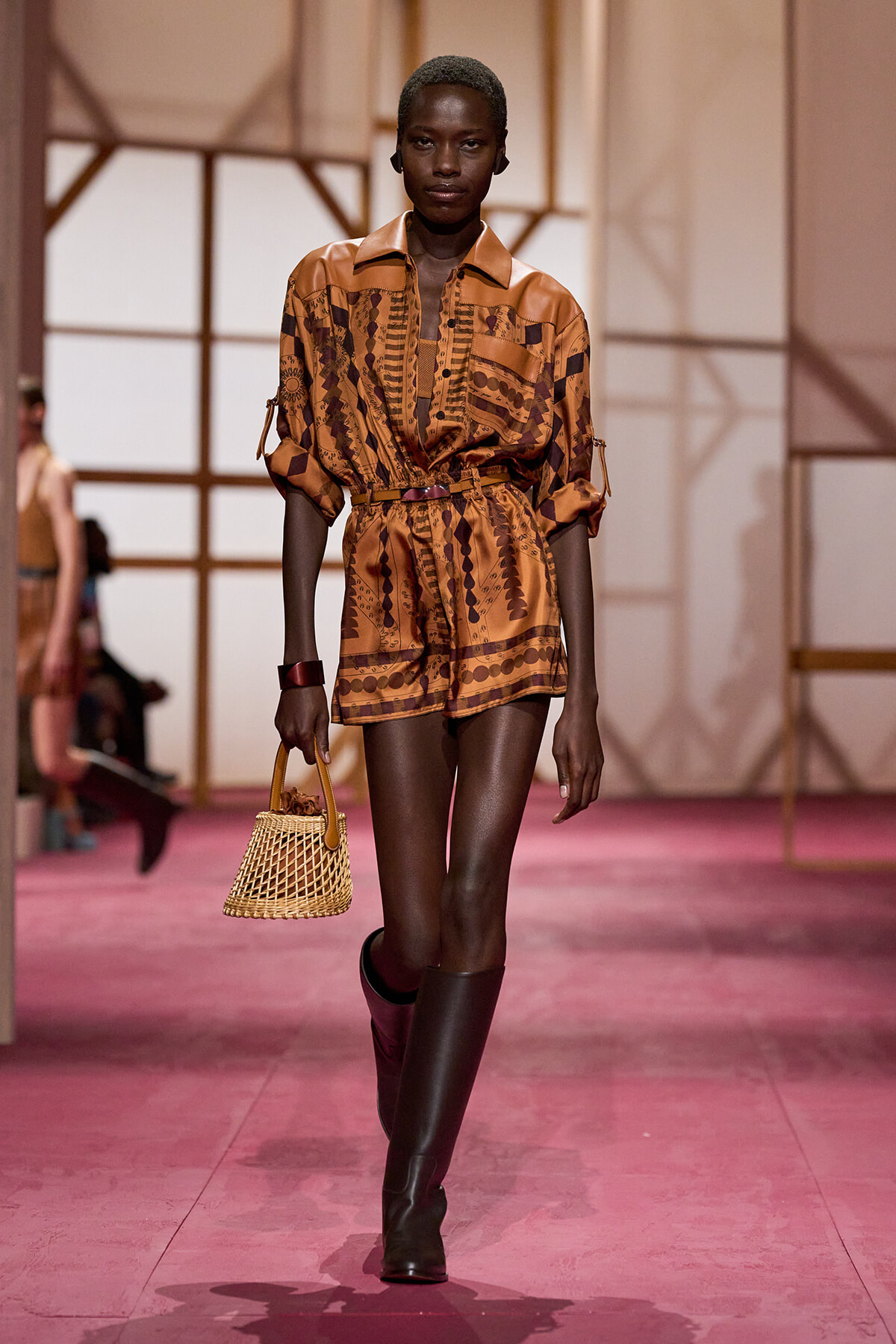 Model walks a fashion runway in a tan-brown patterned romper with a belt, paired with knee-high boots and a small woven wicker handbag