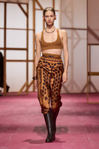 Model on a runway wearing a tan crop top, high-waisted brown patterned pants, and black knee-high boots; geometric print.