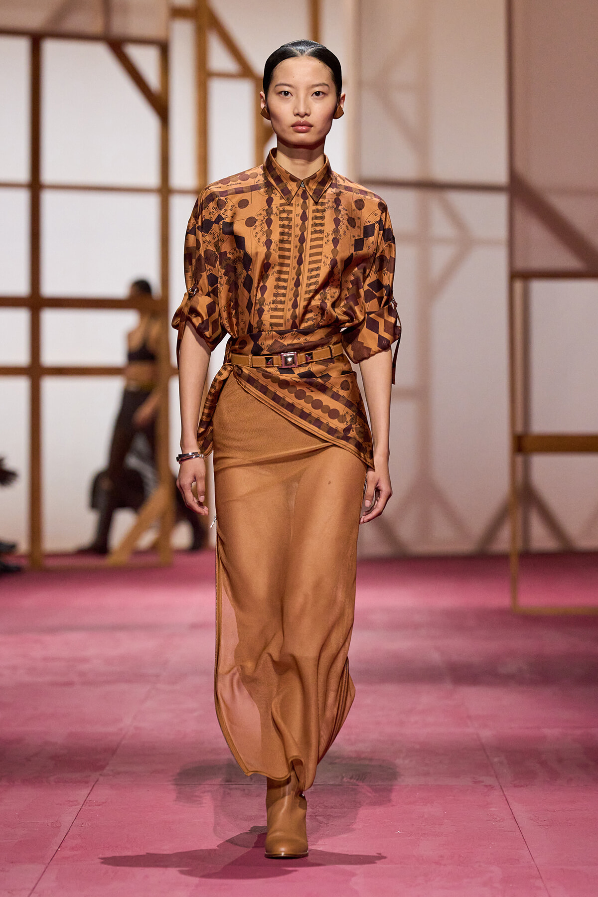 Model walks a fashion runway wearing a brown patterned blouse tucked into a matching wrap skirt with a belt, tan boots, and slicked-back hair.