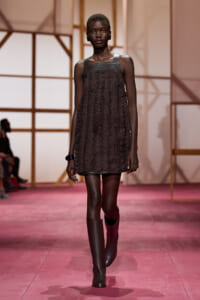 Runway model in a short, textured black dress with a leather bodice and knee-high boots on a pink catwalk.