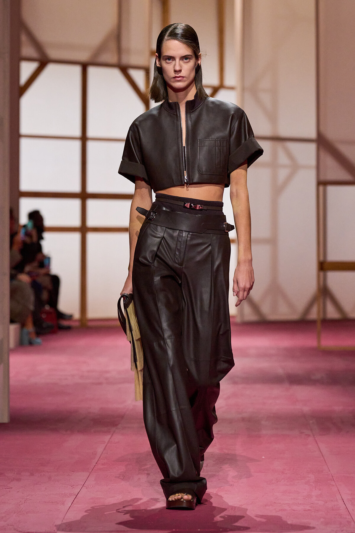 Model walks a pink runway in a black cropped leather jacket with a front zipper and matching high-waisted leather pants.