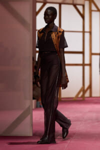 Black model walks the runway in a black zip-front top, orange-brown patterned sleeveless vest, and wide, zipper-adorned pants with black ankle boots.