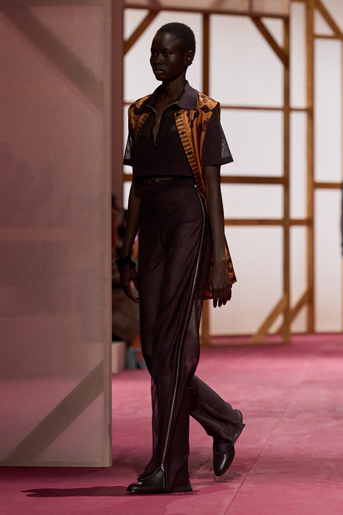 Black model walks the runway in a black zip-front top, orange-brown patterned sleeveless vest, and wide, zipper-adorned pants with black ankle boots.