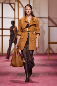 Model on the runway wearing a camel utility trench with belt, rolled sleeves, black crop top, sheer black skirt, knee‑high boots, and a tan tote bag.
