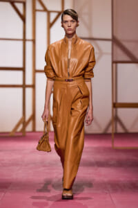 Model on a pink runway wearing a camel leather two-piece: oversized bomber jacket tucked into high-waisted pants, with a small tan handbag