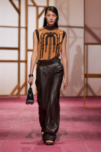 Model on a runway wearing a tan sleeveless top with a black geometric print and high-waisted black leather pants, carrying a small black handbag.