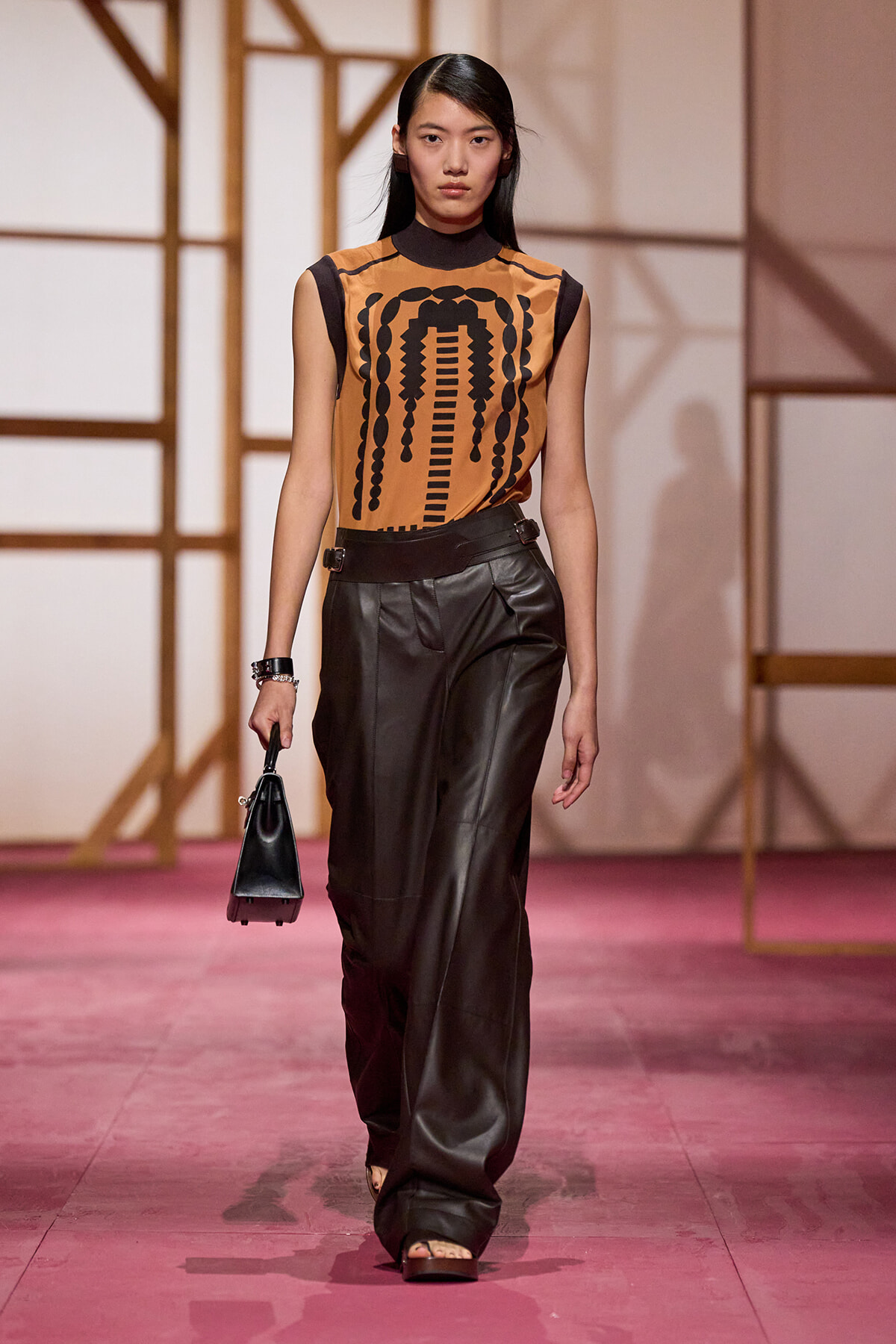 Model on a runway wearing a tan sleeveless top with a black geometric print and high-waisted black leather pants, carrying a small black handbag.