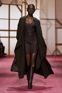 Model walking the runway in a long dark coat over a sheer black dress and tall boots on a pink catwalk.