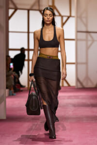 Model walks a fashion runway in a black bra-style top, sheer black midi skirt, mustard belt, tall brown boots, and a dark handbag.