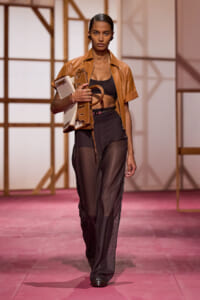 Model walking a fashion runway in a tan cropped leather jacket, black top, sheer black pants, carrying a beige handbag under her arm.