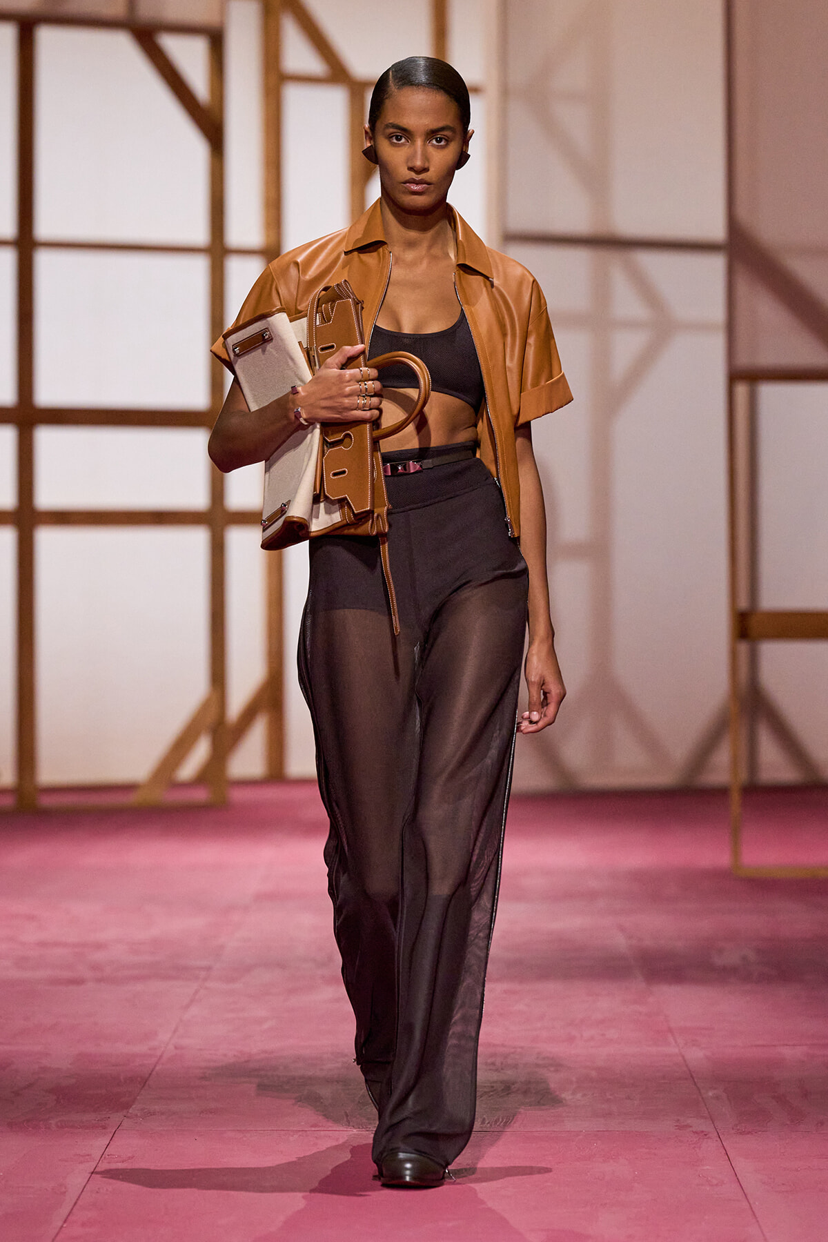 Model walking a fashion runway in a tan cropped leather jacket, black top, sheer black pants, carrying a beige handbag under her arm.
