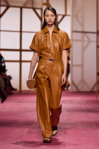 Model walking the runway in a tan leather two-piece outfit with a zip-front top and wide-leg pants, carrying a small round bag.