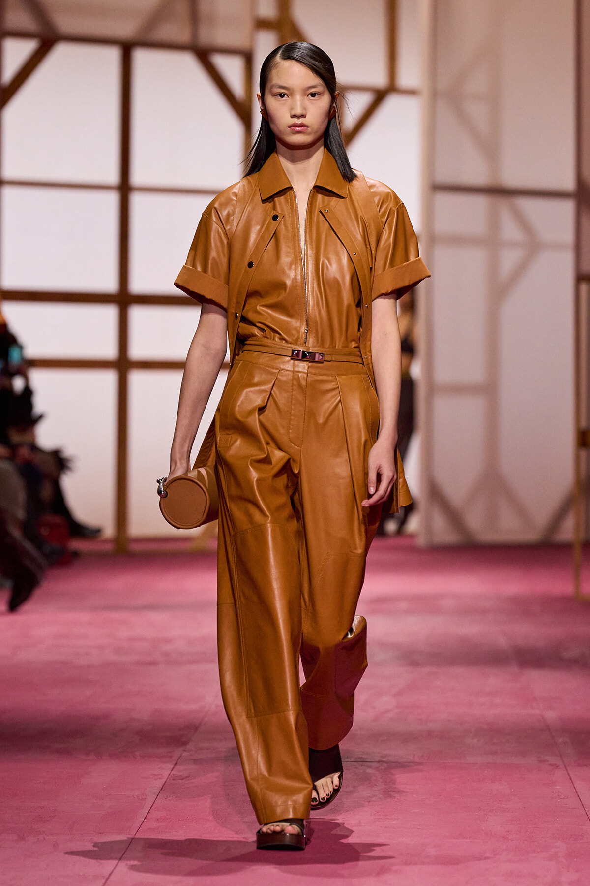 Model walking the runway in a tan leather two-piece outfit with a zip-front top and wide-leg pants, carrying a small round bag.