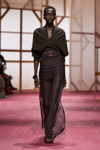 Model walking a fashion runway in a dark draped crop top and sheer black skirt-pants on a pink catwalk.