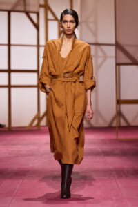 Model walks the runway in a camel-toned belted jumpsuit with rolled sleeves and black ankle boots, against a geometric wooden backdrop.