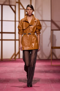 Model walking the runway in a camel leather trench with a belt, paired with sheer black pants and ankle boots.