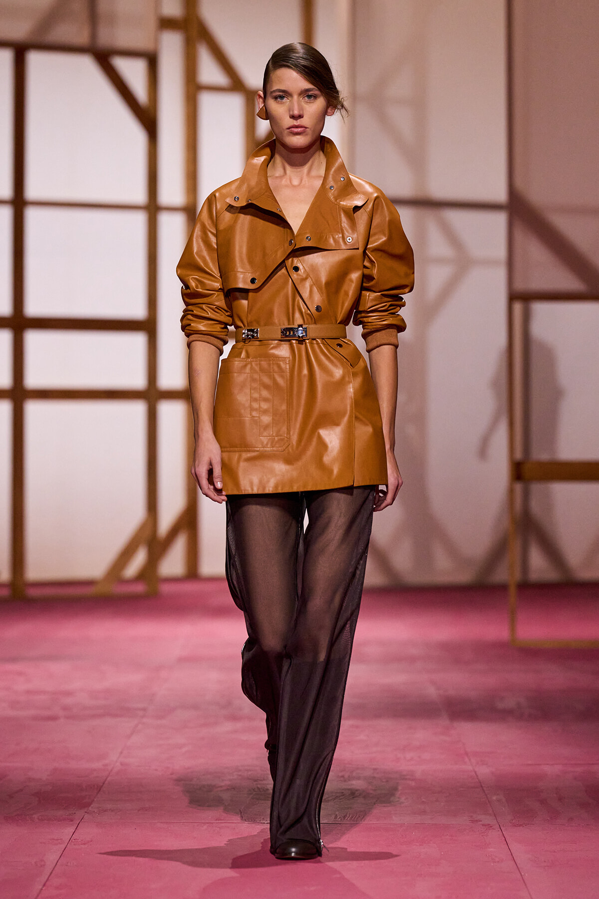 Model walking the runway in a camel leather trench with a belt, paired with sheer black pants and ankle boots.